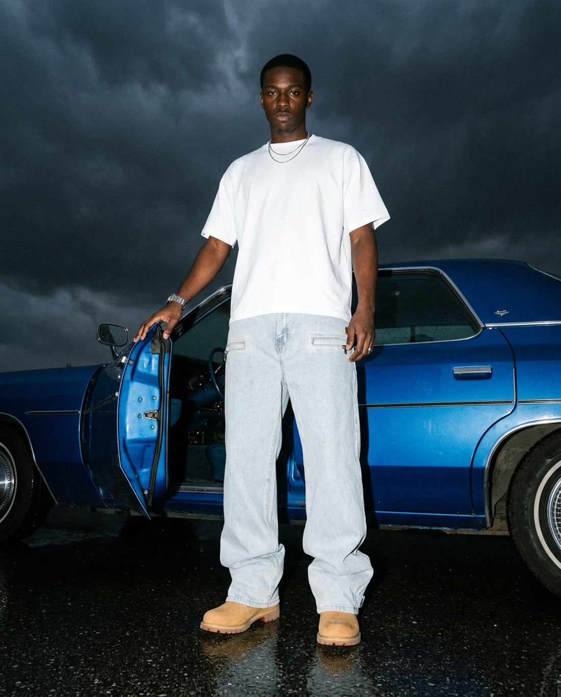 Man in light-wash Omavi baggy jeans beside a blue vintage car under a storm sky